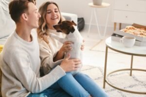 A smiling couple sitting with a small dog between them in a bright living room